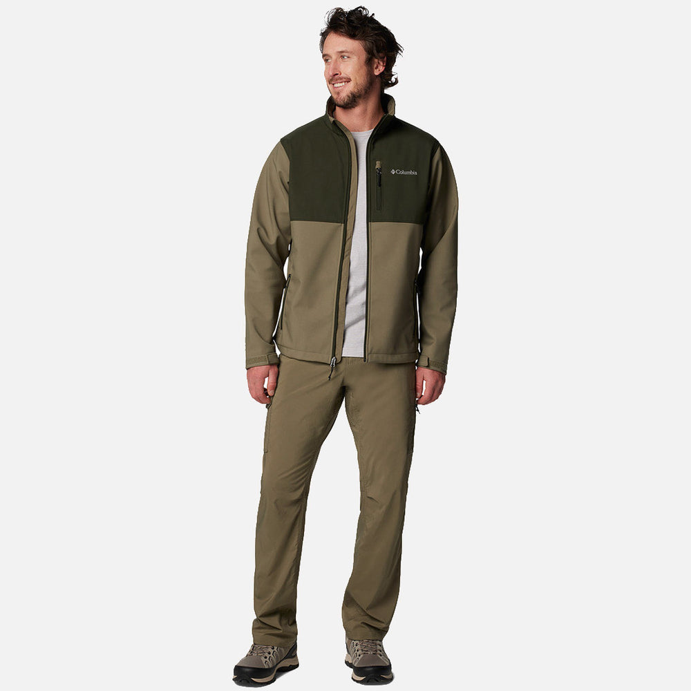 גבר לבוש במעיל Ascender Softshell של ColumbiaIL בצבעי זית וירוק כהה, עם מכנסיים תואמים ונעלי ספורט אפורות, מחייך על רקע לבן חלק.