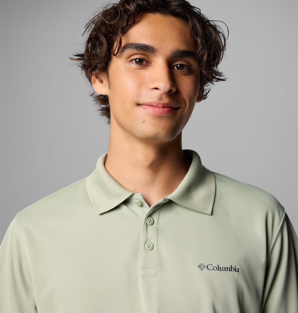 צעיר בעל שיער חום גלי מחייך קלות, לבוש בחולצת פולו בצבע ירוק בהיר של המותג ColumbiaIL Utilizer על רקע אפור אחיד.