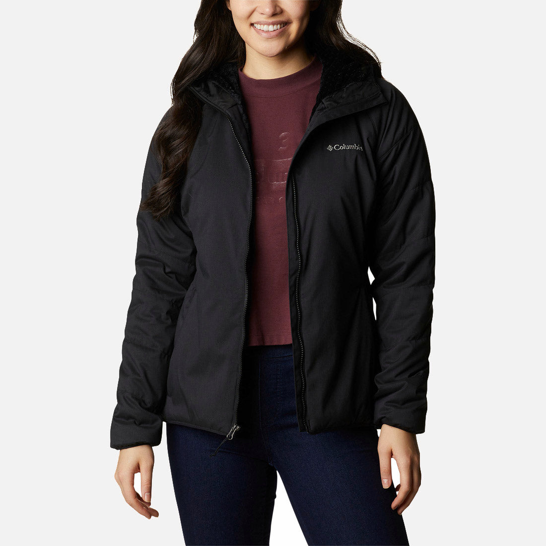 אישה לבושה במעיל Softshell שחור מדגם Kruser Ridge II של ColumbiaIL, מעל חולצה בצבע בורדו וג'ינס כהה, מחייכת בעודה עומדת על רקע לבן חלק.