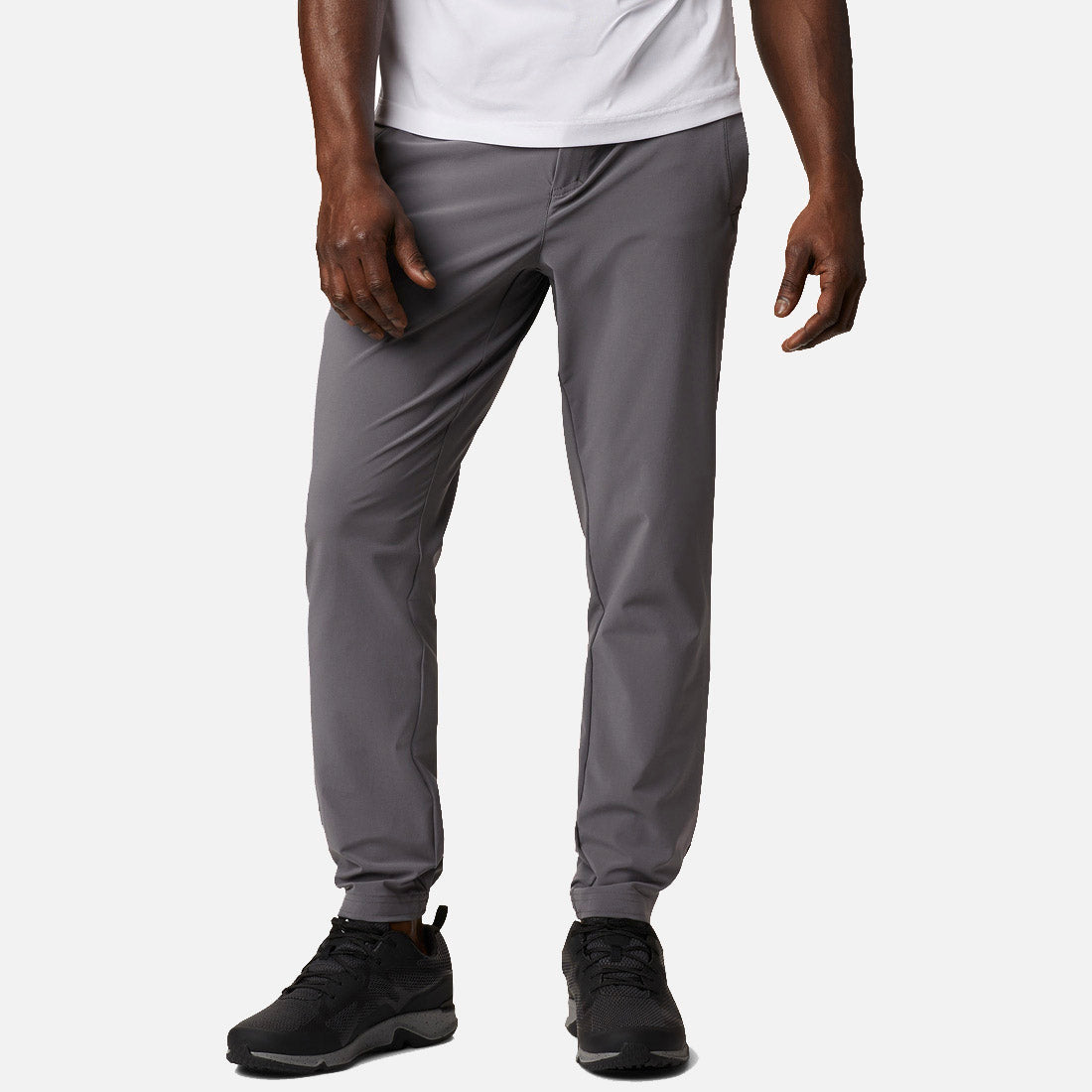 גבר מדגמן את מכנסי ה-Jogger הארוגים של Columbia Lodge מבית ColumbiaIL — מכנסי ריצה אפורים בשילוב עם חולצת טי לבנה ונעלי ספורט שחורות. רק פלג גופו התחתון נראה על רקע לבן אחיד.