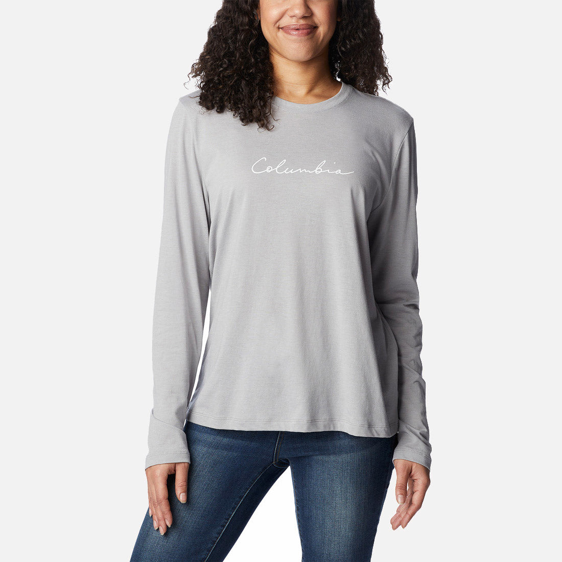 אישה לובשת חולצת Columbia Trek Relaxed L/S T מבית columbiaIL, עם הכיתוב "Columbia" באותיות כתב יד לבנות על החזה, יחד עם ג'ינס כחול כהה, כשהיא עומדת על רקע לבן חלק.