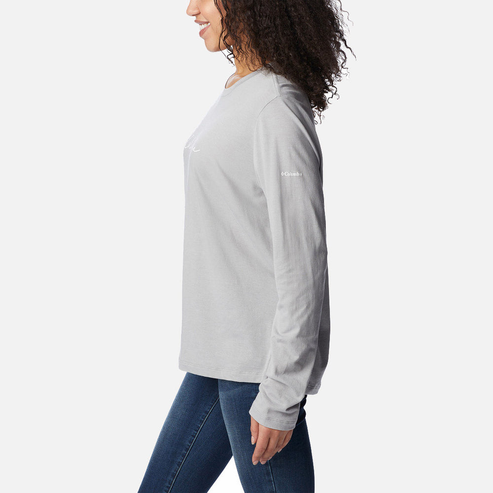 אישה עם שיער מתולתל לובשת חולצת Columbia Trek Relaxed L/S T מבית columbiaIL וג'ינס כחול, עומדת בפרופיל על רקע לבן חלק.
