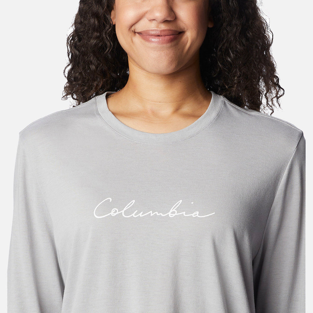 אישה עם שיער מתולתל מחייכת כשהיא לובשת חולצת Columbia Trek Relaxed L/S T מבית columbiaIL, שעליה הכיתוב "Columbia" באותיות כתב יד לבנות על החזה.