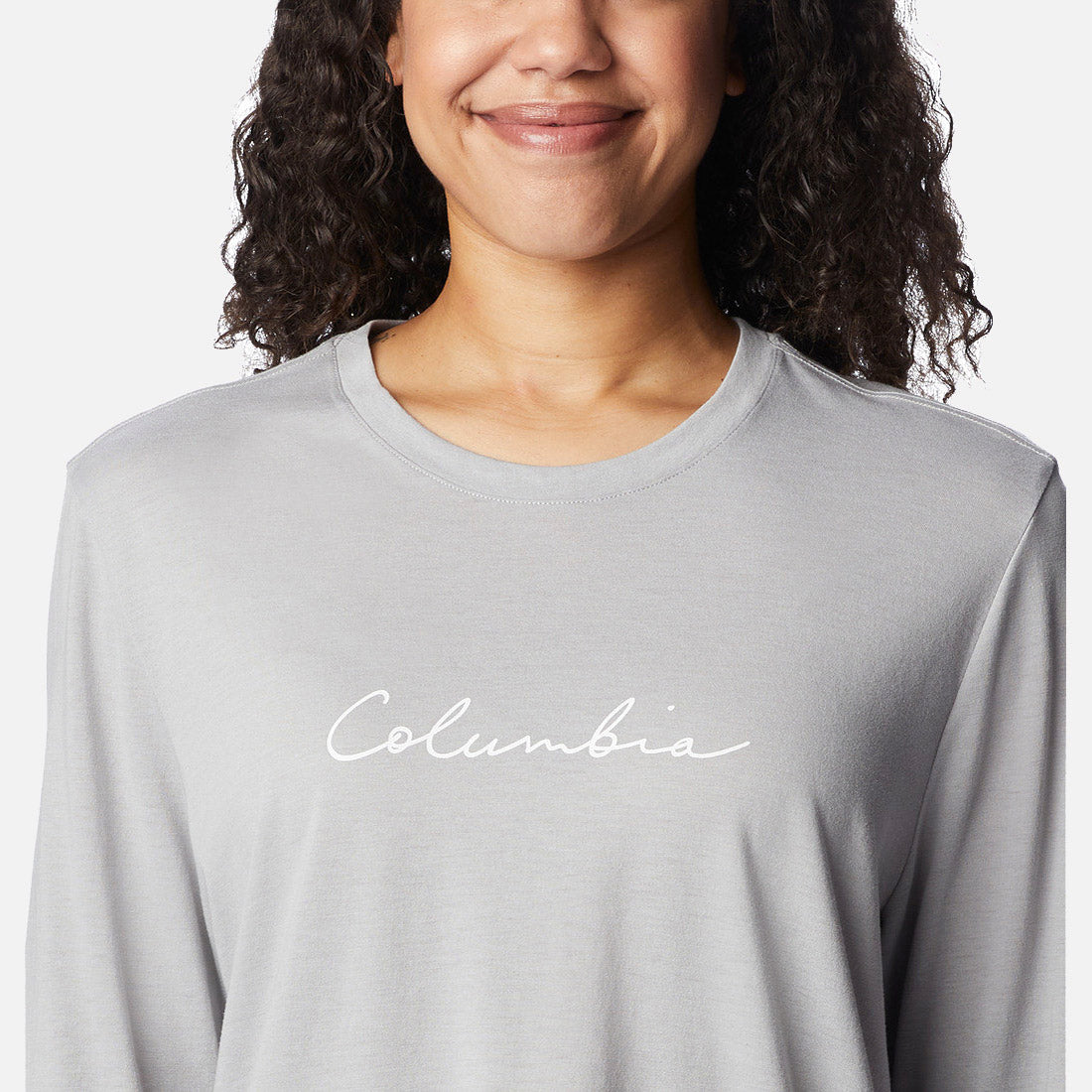 אישה עם שיער מתולתל מחייכת כשהיא לובשת חולצת Columbia Trek Relaxed L/S T מבית columbiaIL, שעליה הכיתוב "Columbia" באותיות כתב יד לבנות על החזה.