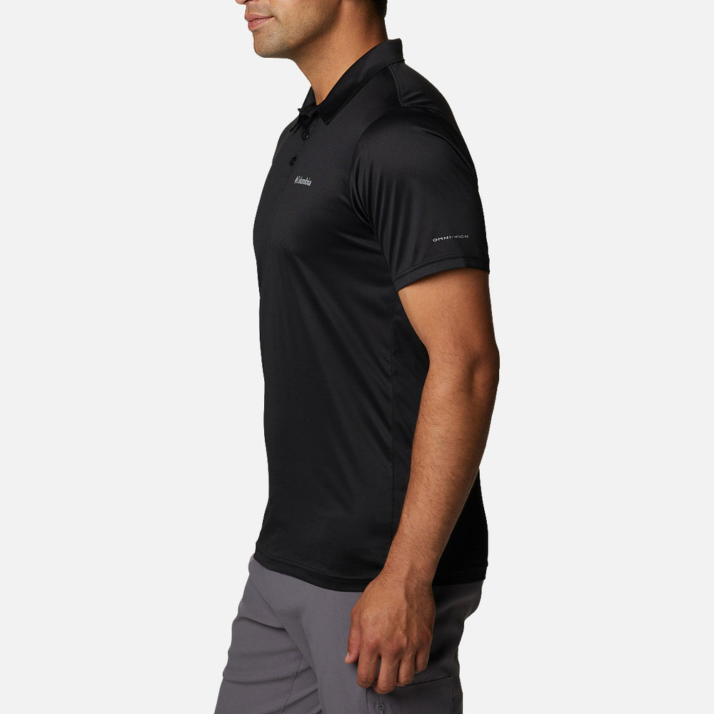 אדם עומד בפרופיל, לבוש בחולצת הפולו "Columbia Hike Polo Black" מבית columbiaIL, בשילוב עם מכנסיים אפורים על רקע לבן חלק. על החולצה מוטבעים לוגואים עדינים על החזה ועל השרוול.