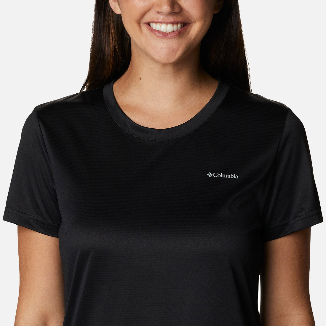 אישה מחייכת למצלמה כשהיא לובשת חולצת Columbia Hike S/S Crew Black של קולומביה. התמונה, שנחתכה בגובה הכתפיים, מדגישה את הצווארון העגול ואת הלוגו הקטן של קולומביה בצד שמאל של החזה.
