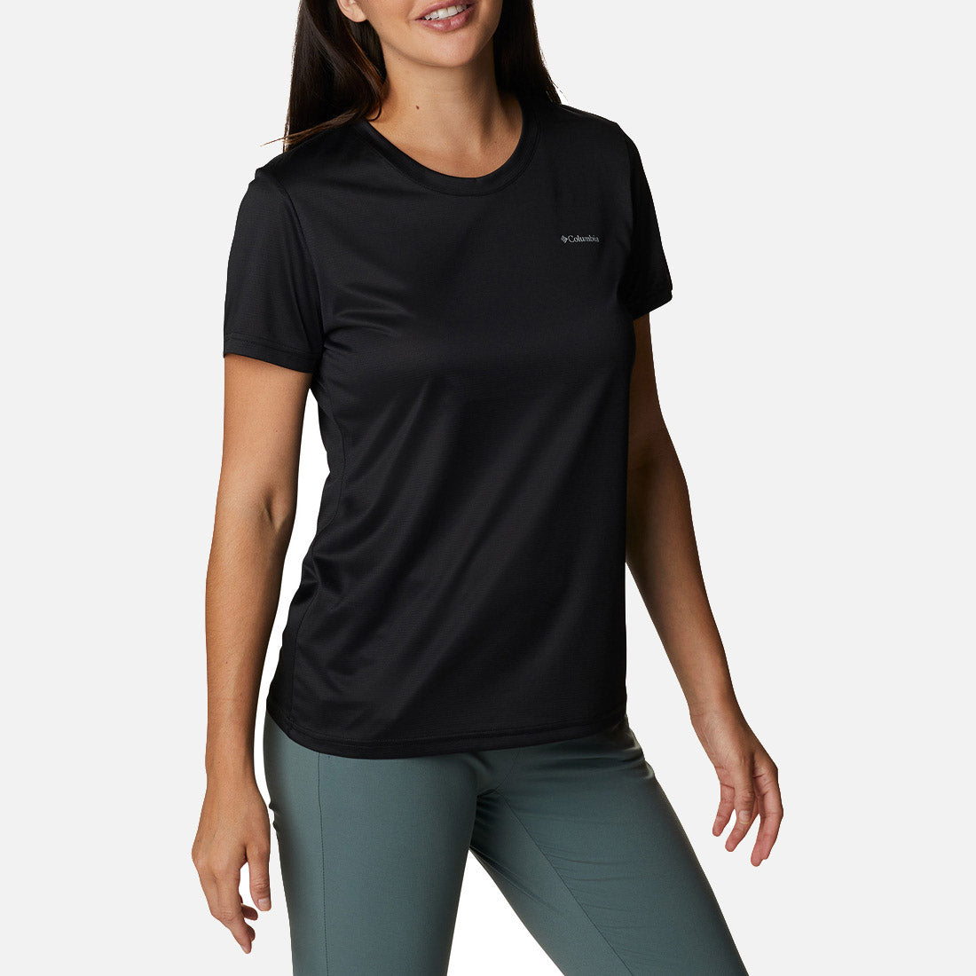 אדם לבוש בחולצת Columbia Hike S/S Crew שחורה של ColumbiaIL ובמכנסיים ירוקים עומד על רקע לבן, מחייך, כשידו האחת רפויה והשנייה כפופה קלות.