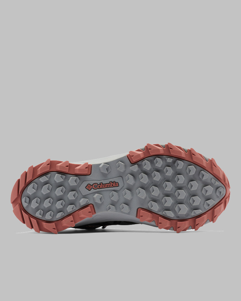 בתמונה נראית הסוליה של נעל Columbia Peakfreak II Mid Outdry W. הסוליה כוללת דפוס משושה ומשולש בצבע אפור עם קצוות בצבע חום-אדמדם לשיפור האחיזה, ולוגו אדום של Columbia במרכזה. הרקע הוא אפור בהיר.