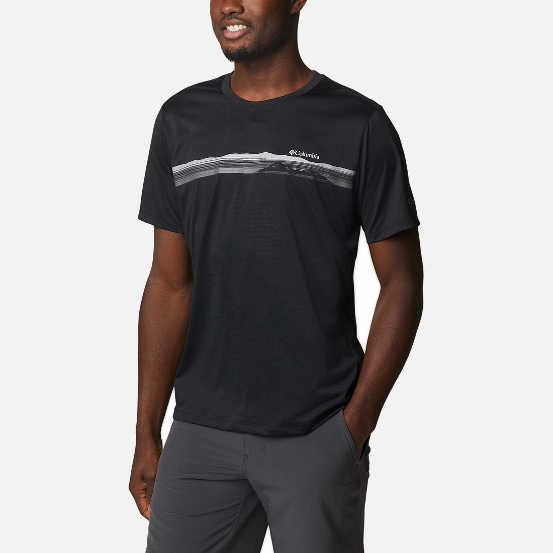 גבר לובש את חולצת ה-Columbia Hike Graphic S/S T מבית columbiaIL, חולצת טי שחורה עם שרוולים קצרים ופסים דקים בצבע אפור בהיר, בשילוב עם מכנסיים אפורים. הוא עומד על רקע לבן, ידיו בכיסים וחיוך על פניו — מראה מושלם להרפתקאות בטבע.