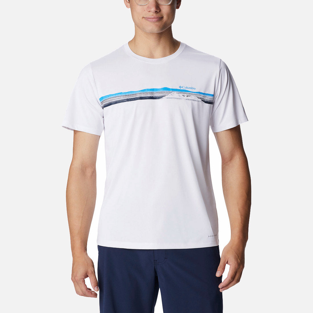 גבר לבוש בחולצת ה-Hike Graphic S/S T של קולומביה (columbiaIL) — בצבע לבן עם פסים כחולים ושחורים על החזה — ובמכנסיים קצרים בצבע כחול כהה, עומד על רקע לבן חלק; לבוש אידיאלי לפעילויות בחוץ בימי הקיץ החמים.
