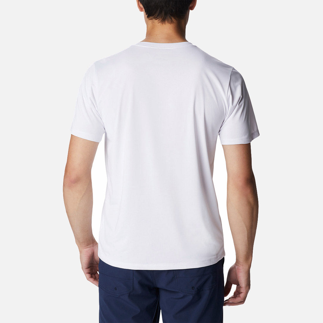 נראה גבר מאחור, לבוש בחולצת Columbia Hike Graphic S/S T מבית columbiaIL ובמכנסיים כחולים כהים, עומד על רקע בהיר וחלק.
