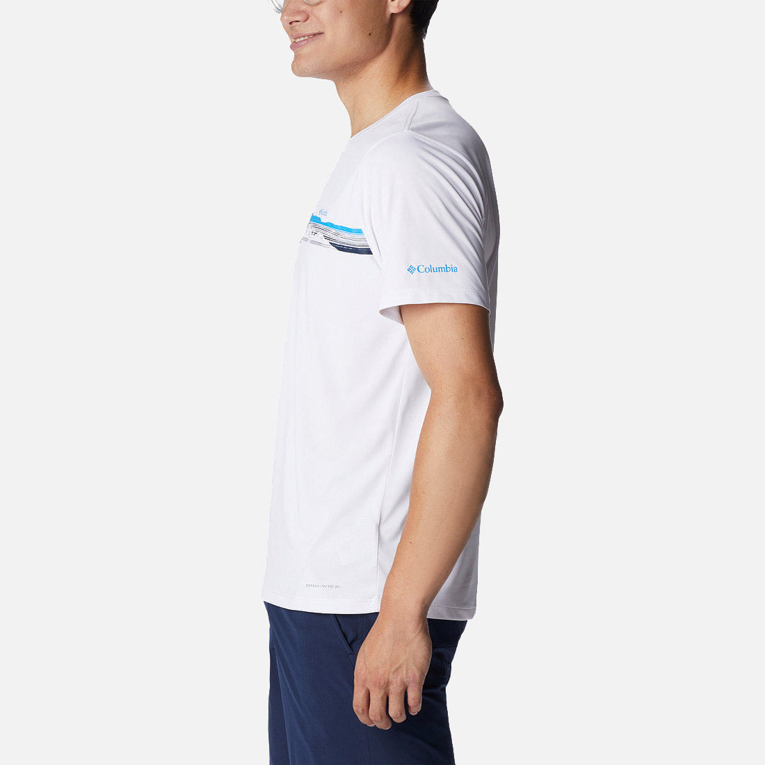אדם לבוש בחולצת ה-Hike Graphic S/S T של קולומביה, בעיצוב פסים כחולים ושחורים, ומכנסיים קצרים בצבע כחול כהה, מצולם מהצד על רקע לבן חלק.