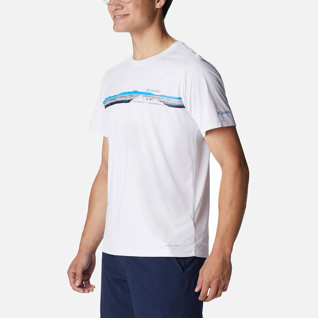 גבר לבוש בחולצת טי-שירט Columbia Hike Graphic S/S T של ColumbiaIL, עם הדפס הרים בכחול ושחור על רקע לבן, בשילוב מכנסיים קצרים בצבע כחול כהה — אידיאלי לחובבי פעילויות בחיק הטבע — ניצב על רקע לבן חלק.