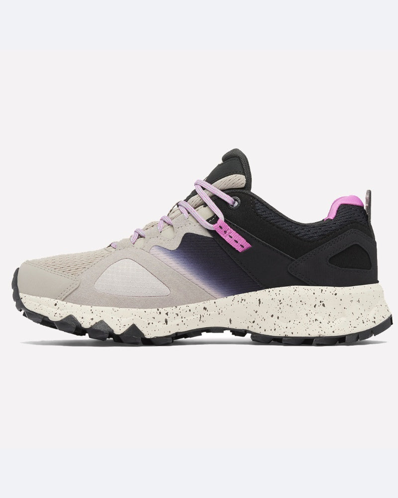 ה-Peakfreak Hera Outdry של ColumbiaIL היא נעל ריצת שטח עם קצה בהיר, עקב שחור, חלק אמצעי בגווני סגול, שרוכים בצבע לילך, סוליה לבנה מנוקדת ואלמנטים סגולים — המשלבת ביצועים בשטח עם סגנון בהשראת HERA.