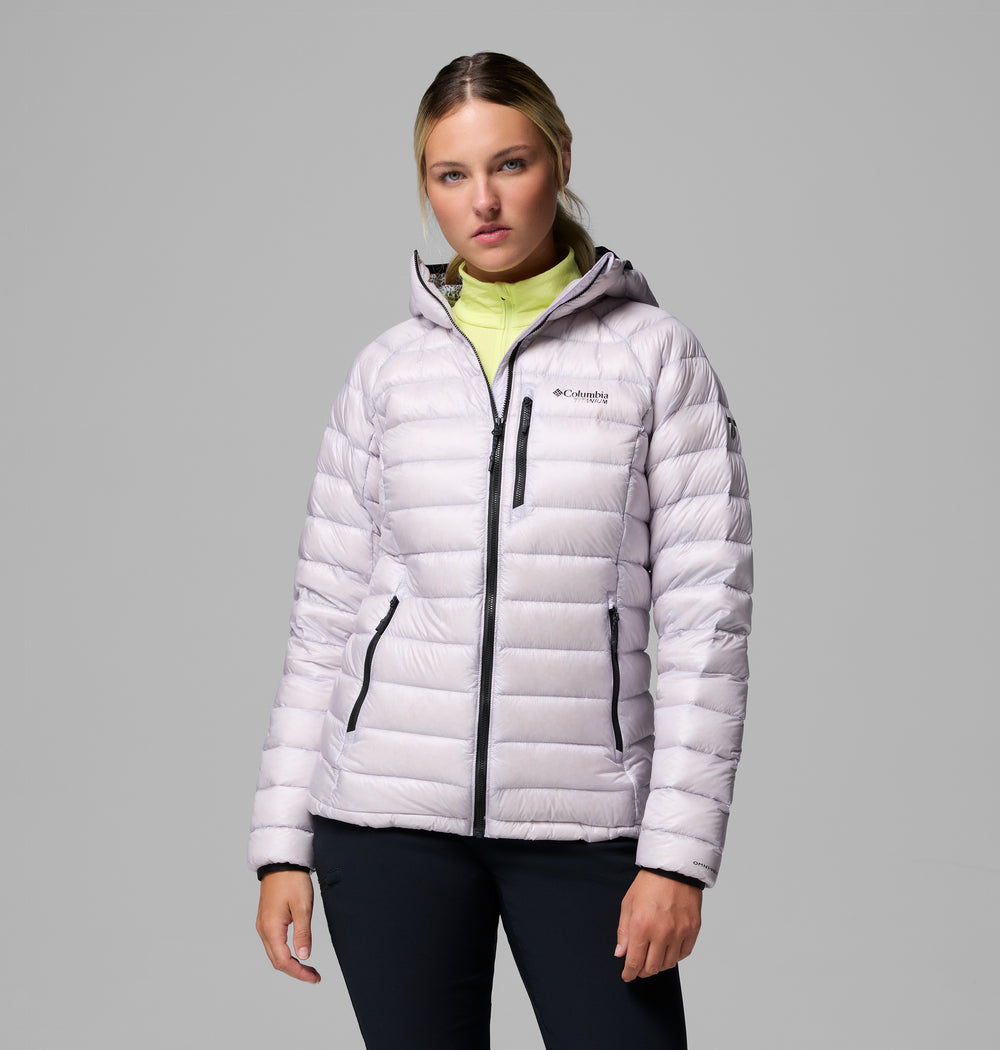 אישה לבושה במעיל פוך עם ברדס מדגם Arctic Crest של המותג ColumbiaIL, עם רוכסנים שחורים, עומדת על רקע אפור אחיד ומביטה במצלמה במבט ניטרלי.