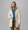 אישה הלובשת את מעיל החורף "Joy Peak II Mid Jacket" של קולומביה IL — מעיל חורף מרופד בצבע בז' בהיר עם ברדס — עומדת בביטחון, לבושה בחולצה כחולה ומכנסיים בצבע חום בהיר, על רקע אפור.