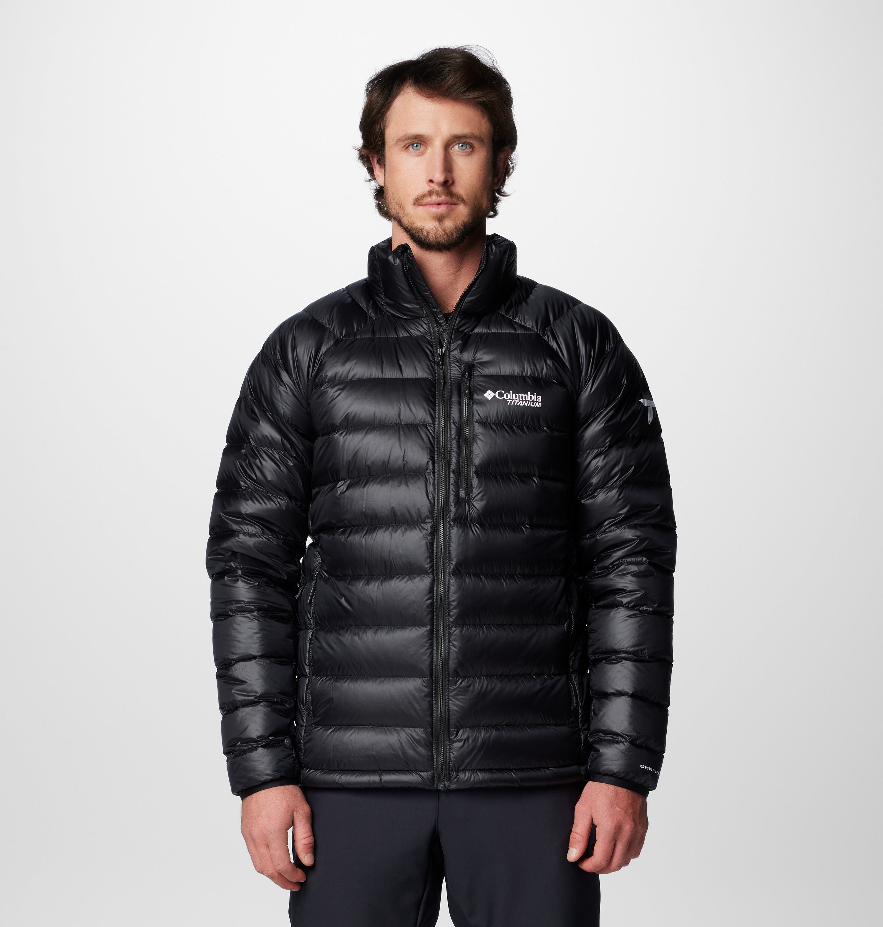 גבר בעל שיער כהה וזקן קצר לובש מעיל פוך Arctic Crest של המותג ColumbiaIL בצבע שחור, עם לוגו המותג על החזה, על רקע אפור בהיר חלק.
