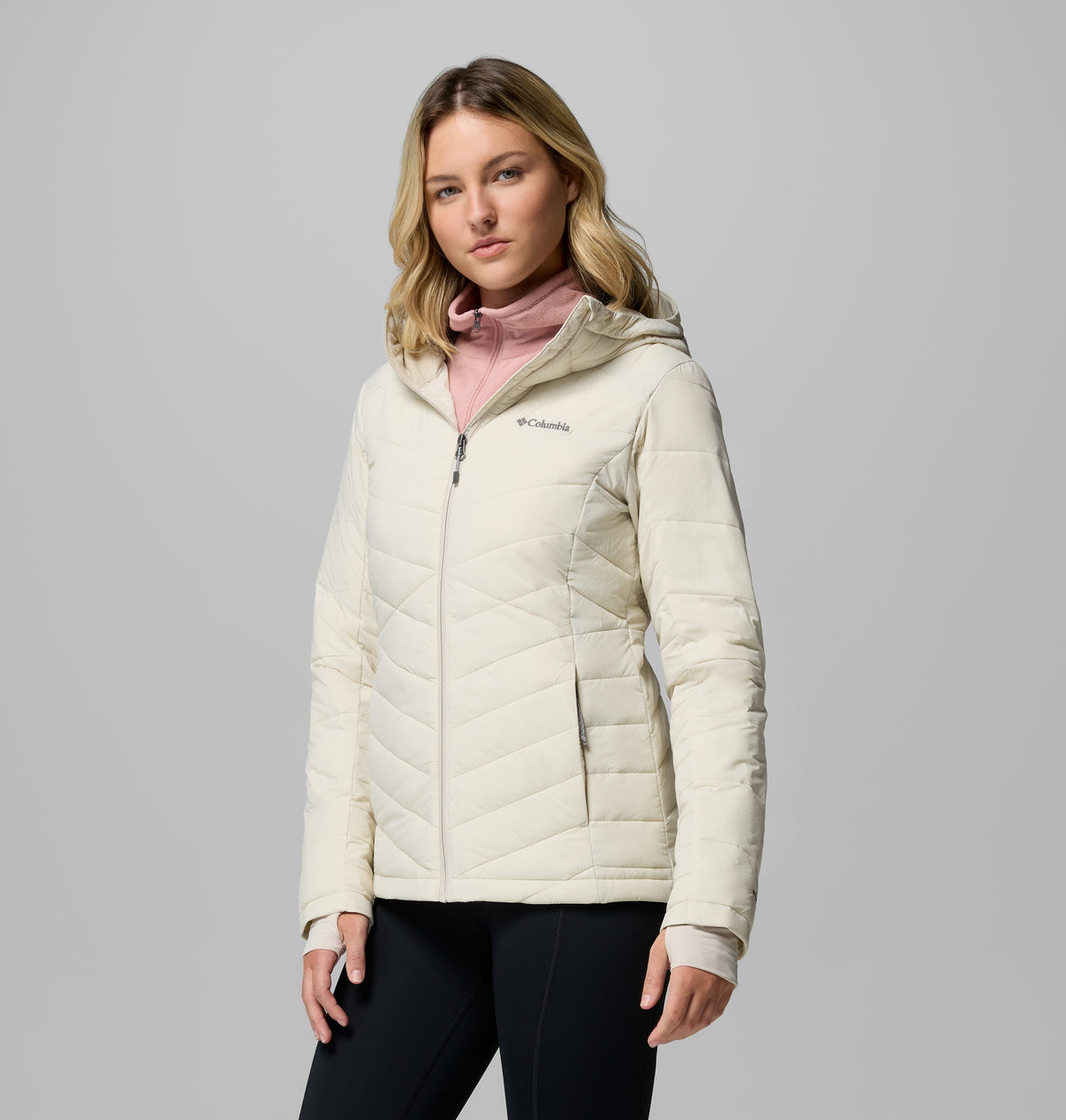 אישה מדגמנת את מעיל ה-Joy Peak II Hooded Jacket של קולומביה ישראל בגוון בהיר, מעל חולצת גולף ורודה בהירה, כשהיא עומדת על רקע אפור עם הבעה ניטרלית, ומדגישה את סגנון הלבוש האופנתי לפעילויות בחוץ.