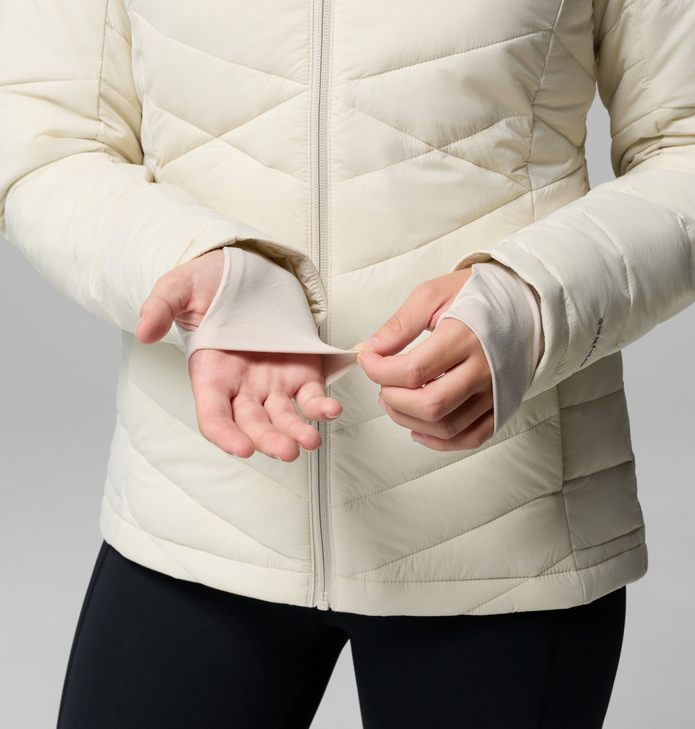 הדוגמנית לובשת את מעיל ה-Joy Peak II Hooded Jacket של קולומביה ישראל ומכנסיים שחורים, ומדגימה את חור האגודל בשרוול כשהיא מושיטה את ידיה קדימה על רקע אחיד — דרך אידיאלית להבליט את המעיל הנעים הזה המיועד לפעילויות בחוץ.