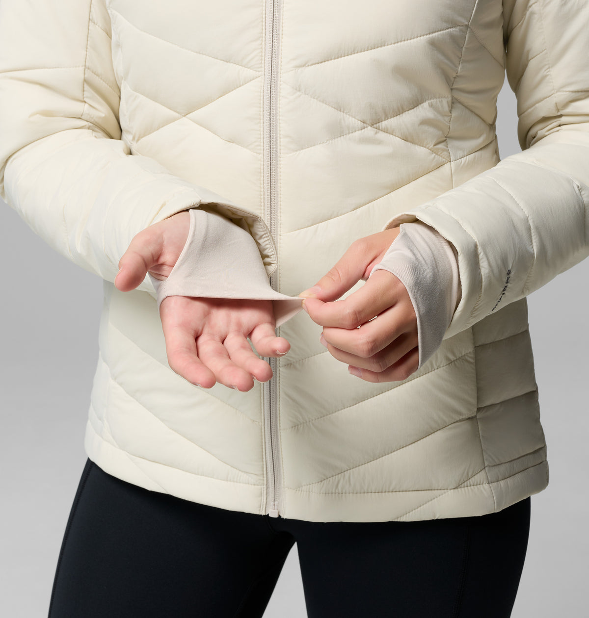 הדוגמנית לובשת את מעיל ה-Joy Peak II Hooded Jacket של קולומביה ישראל ומכנסיים שחורים, ומדגימה את חור האגודל בשרוול כשהיא מושיטה את ידיה קדימה על רקע אחיד — דרך אידיאלית להבליט את המעיל הנעים הזה המיועד לפעילויות בחוץ.