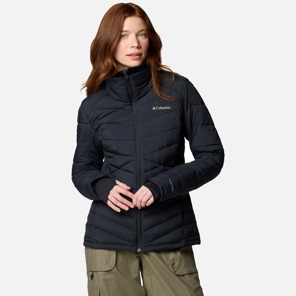 אישה עם שיער חום ארוך לובשת מעיל שחור מדגם "Joy Peak II Full Zip" של המותג "ColumbiaIL" ומכנסי קרגו בצבע ירוק זית, ועומדת על רקע אפור בהיר אחיד.