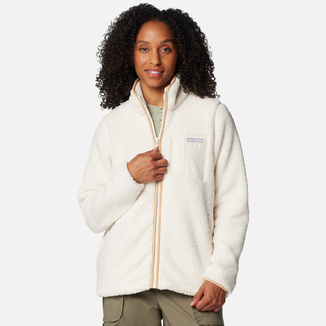 אדם בעל שיער מתולתל לובש מעיל פליס מסוג ColumbiaIL West Bend Full Zip II בצבע שמנת, עם כיס בחזה, ומכנסיים בצבע ירוק זית, והוא עומד על רקע בהיר וחלק.