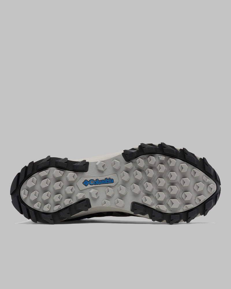 בתמונה נראית הסוליה של מגף הטיולים לגברים Peakfreak II Mid Outdry M מבית columbiaIL, הכוללת בסיס אפור, קצוות שחורים, דפוס משושה ולוגו כחול של Columbia במרכז. הרקע הוא אפור אחיד.