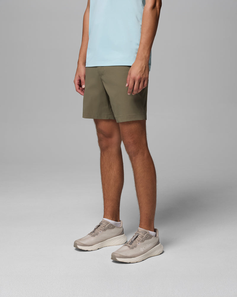 אדם לבוש בחולצה תכלת, מכנסיים קצרים מדגם "Tech Trail Utility Short" של קולומביה ישראל בצבע ירוק זית, ונעלי ספורט בצבע בז', עומד על רקע אפור בהיר אחיד, כשפניו אינן נראות.
