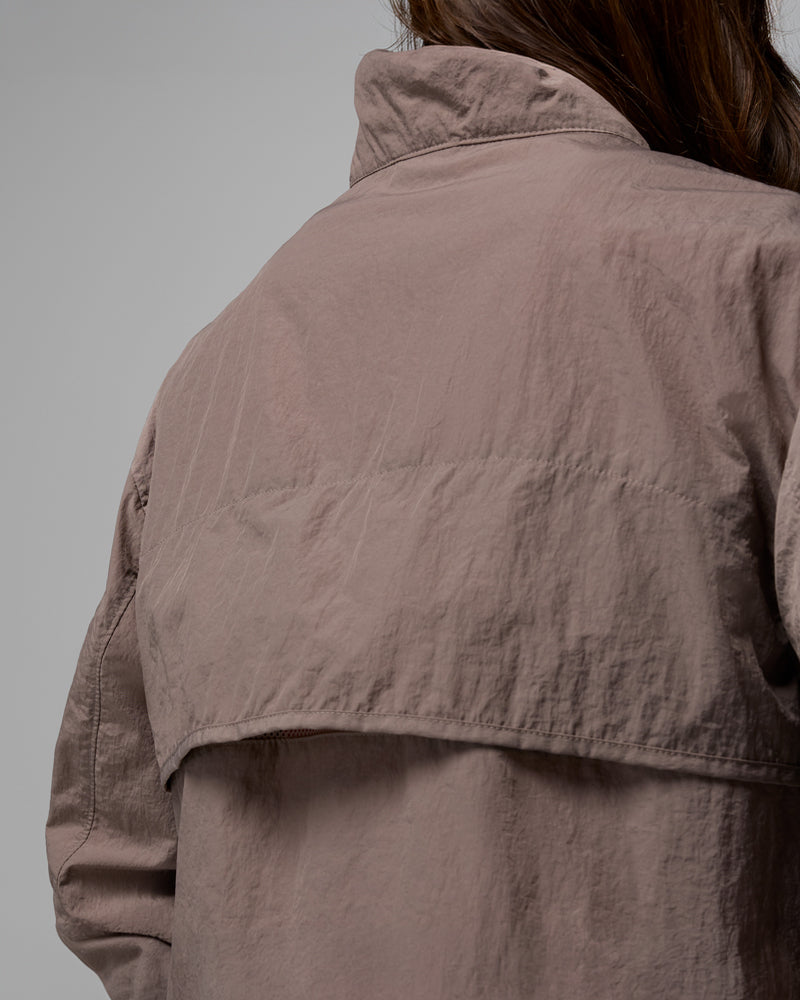 תקריב של אדם הלובש את מעיל הרוח הקצר מסוג "Paracutie Cropped Cargo" של קולומביה ישראל בצבע חום בהיר, הנראה מאחור עם שיער כהה המשתלשל מעל הצווארון על רקע אפור אחיד.