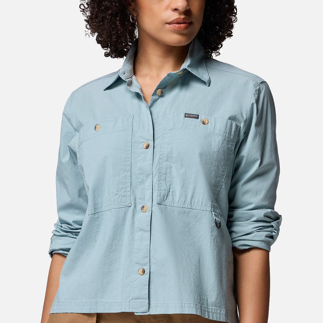 אישה עם שיער מתולתל לובשת את חולצת "Brea Falls Woven Long Sleeve Shirt" של קולומביה ישראל — חולצה קז'ואלית בצבע תכלת עם שני כיסים בחזה ושרוולים מגולגלים — כשהיא עומדת על רקע לבן חלק.