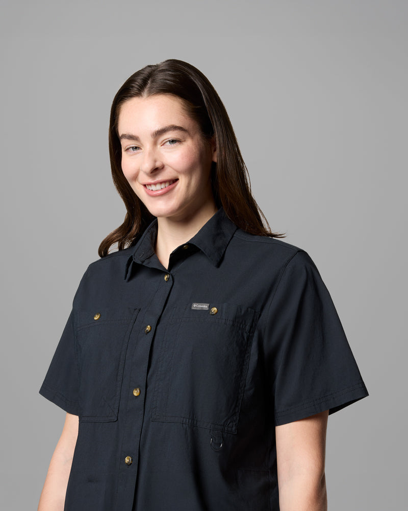 אישה עם שיער חום ארוך לובשת את חולצת ה-Brea Falls Woven S/S של קולומביה ישראל ומחייכת למצלמה על רקע אפור אחיד.