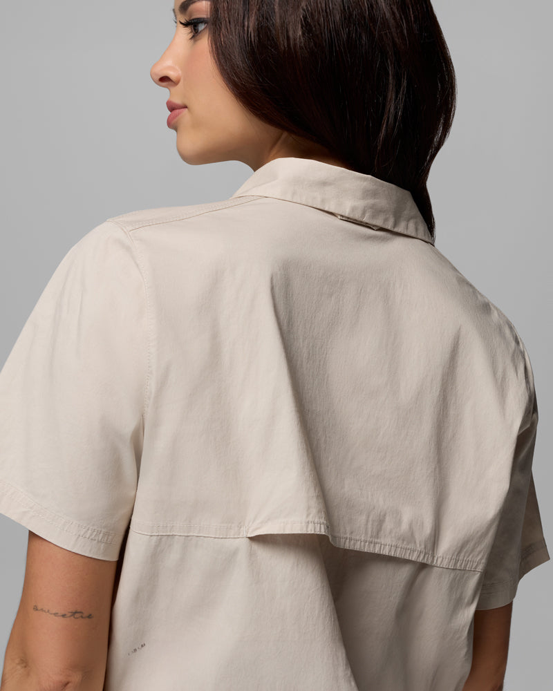 אישה בעלת שיער ארוך וכהה מדגמנת את חולצת ה-Brea Falls Woven S/S של קולומביה ישראל בצבע בז' בהיר; התמונה מוצגת מהצד כדי להדגיש את הפרטים בגב ואת הקעקוע הקטן בסגנון כתב יד על אמת ידה, על רקע אפור בהיר.