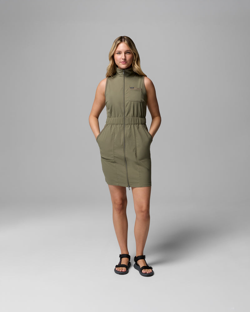 אישה עומדת על רקע אפור אחיד, לבושה בשמלת "Leslie Falls Dress II" של קולומביה ישראל בצבע ירוק זית. השמלה ללא שרוולים, הנסגרת ברוכסן, כוללת חגורה אלסטית במותניים וכיסים. היא משלבת אותה עם סנדלים שחורים ומחייכת קלות למצלמה.