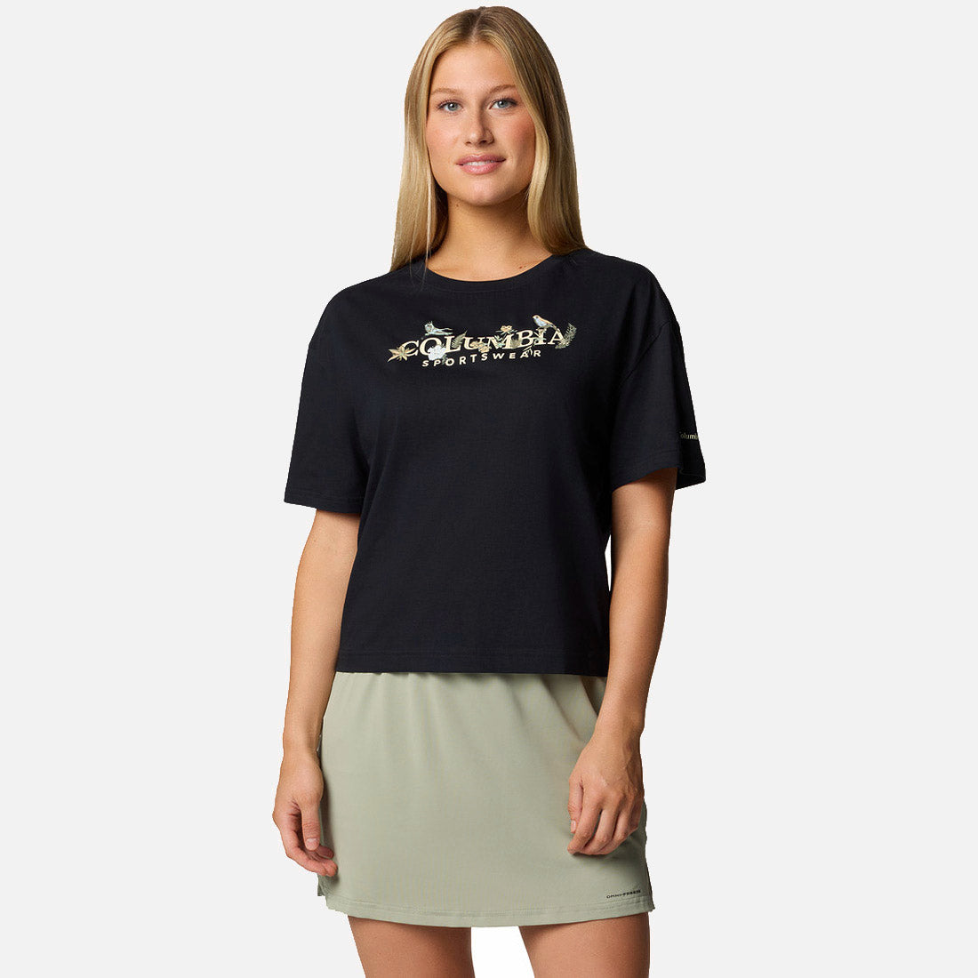 אישה מחייכת, לבושה בחולצת קולומביה IL Graphic Relaxed Crop קצרת שרוולים בצבע שחור וחצאית בצבע ירוק בהיר, עומדת על רקע לבן חלק.