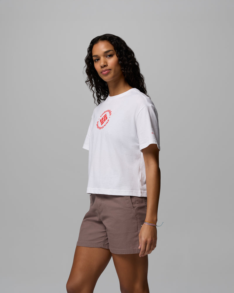 אדם בעל שיער ארוך ומתולתל לובש את חולצת ה-Graphic Relaxed Crop Short Sleeve Shirt של קולומביה ישראל, המעוטרת בעיצוב מעגלי אדום, בשילוב עם מכנסיים קצרים חומים בהירים, על רקע אפור אחיד.