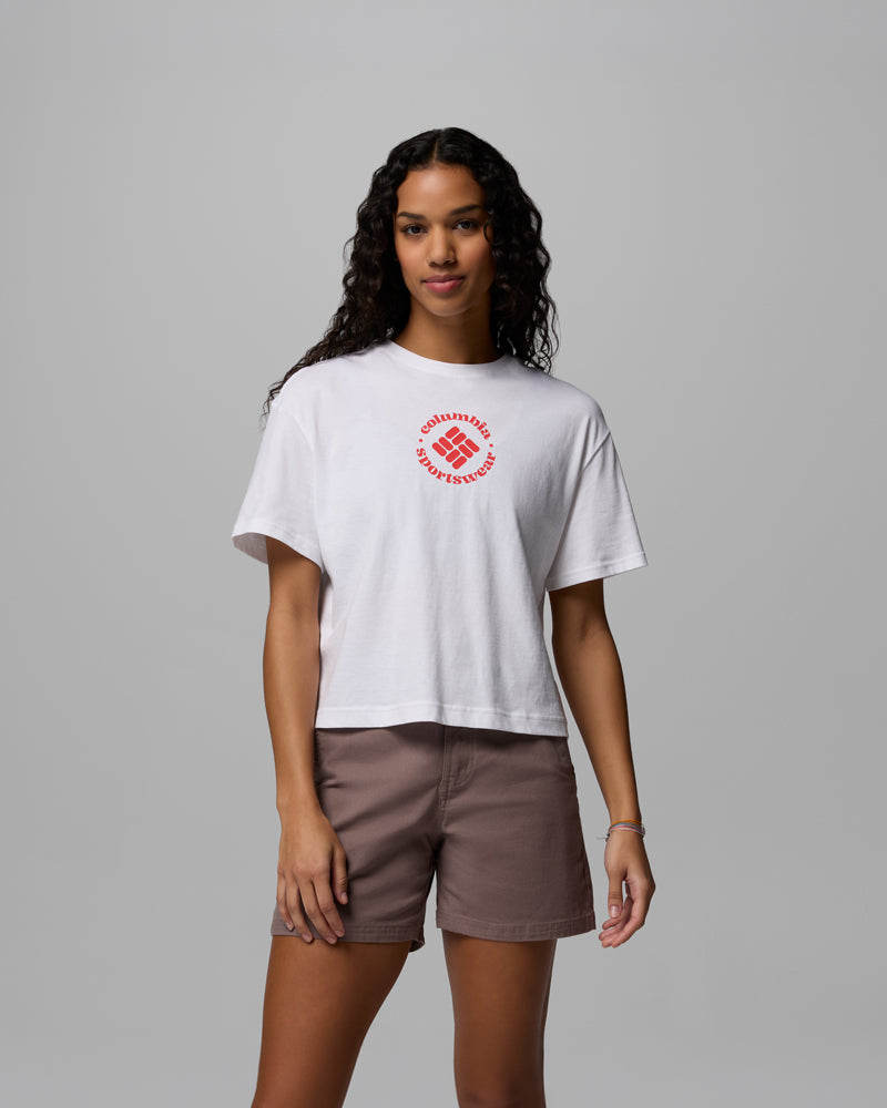 אישה עם שיער ארוך ומתולתל לובשת את חולצת ה-Graphic Relaxed Crop Short Sleeve Shirt של קולומביה ישראל, המעוטרת בעיצוב מעגלי אדום, עם מכנסיים קצרים חומים על רקע אפור אחיד.