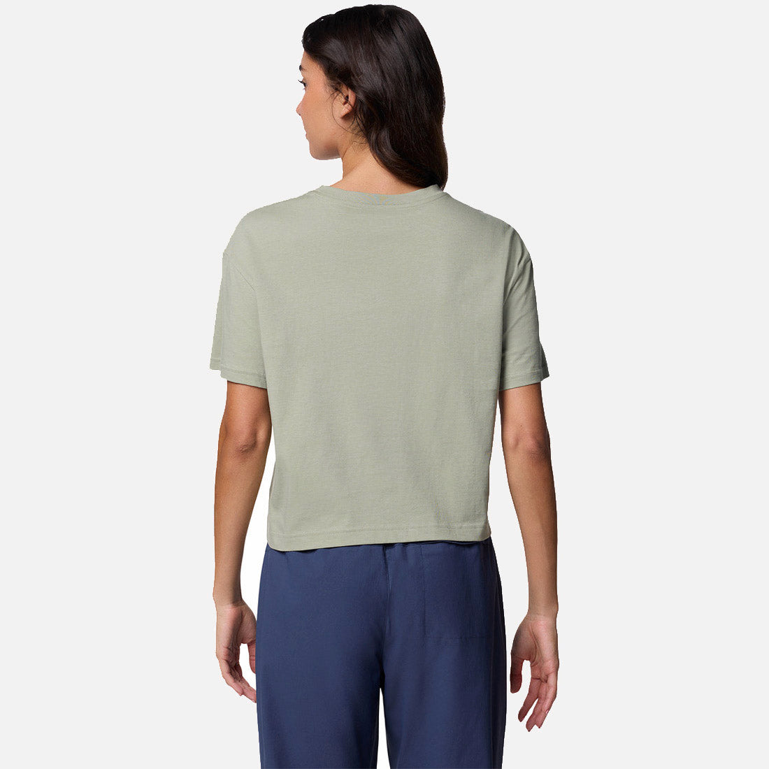 אישה עומדת עם גבה למצלמה, לבושה בחולצת "Graphic Relaxed Crop" קצרת שרוולים של ColumbiaIL בצבע ירוק בהיר ומכנסיים בכחול כהה, על רקע לבן חלק.