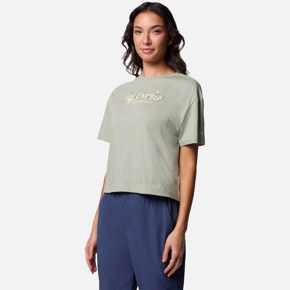 אישה בעלת שיער חום ארוך מדגמנת את חולצת ה-Graphic Relaxed Crop Short Sleeve של ColumbiaIL בצבע ירוק בהיר עם הדפס על החזה, בשילוב עם מכנסיים בצבע כחול כהה על רקע לבן חלק.
