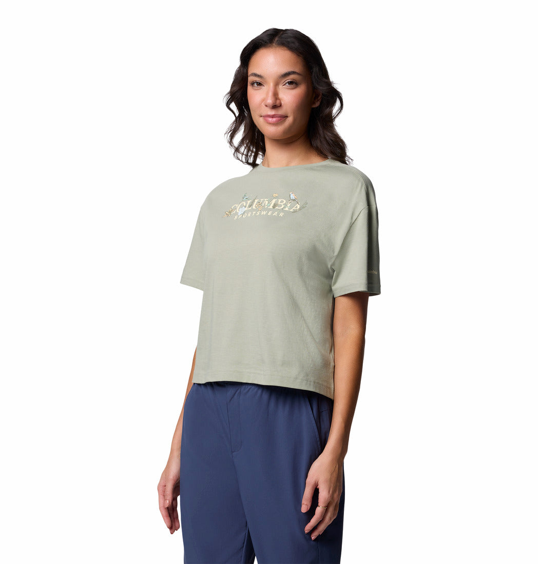 אישה עם שיער חום גלי לובשת את חולצת ה-Graphic Relaxed Crop Short Sleeve Shirt של columbiaIL, בצבע ירוק בהיר עם הדפס פרחוני, בשילוב עם מכנסיים כחולים, כשהיא עומדת על רקע לבן.