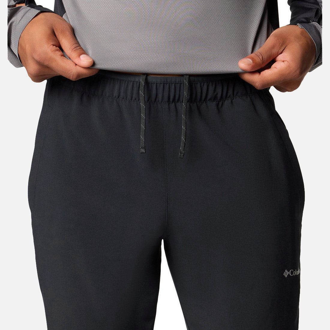 אדם מתאים את חגורת המכנסיים הספורטיביים "Three Pitch Pant" של ColumbiaIL בצבע שחור, בשילוב עם חולצה ארוכת שרוולים בצבע אפור בהיר. בתמונה נראים פלג הגוף העליון והרגליים העליונות, מה שמדגיש את הסגנון הנוח של בגדי הספורט.