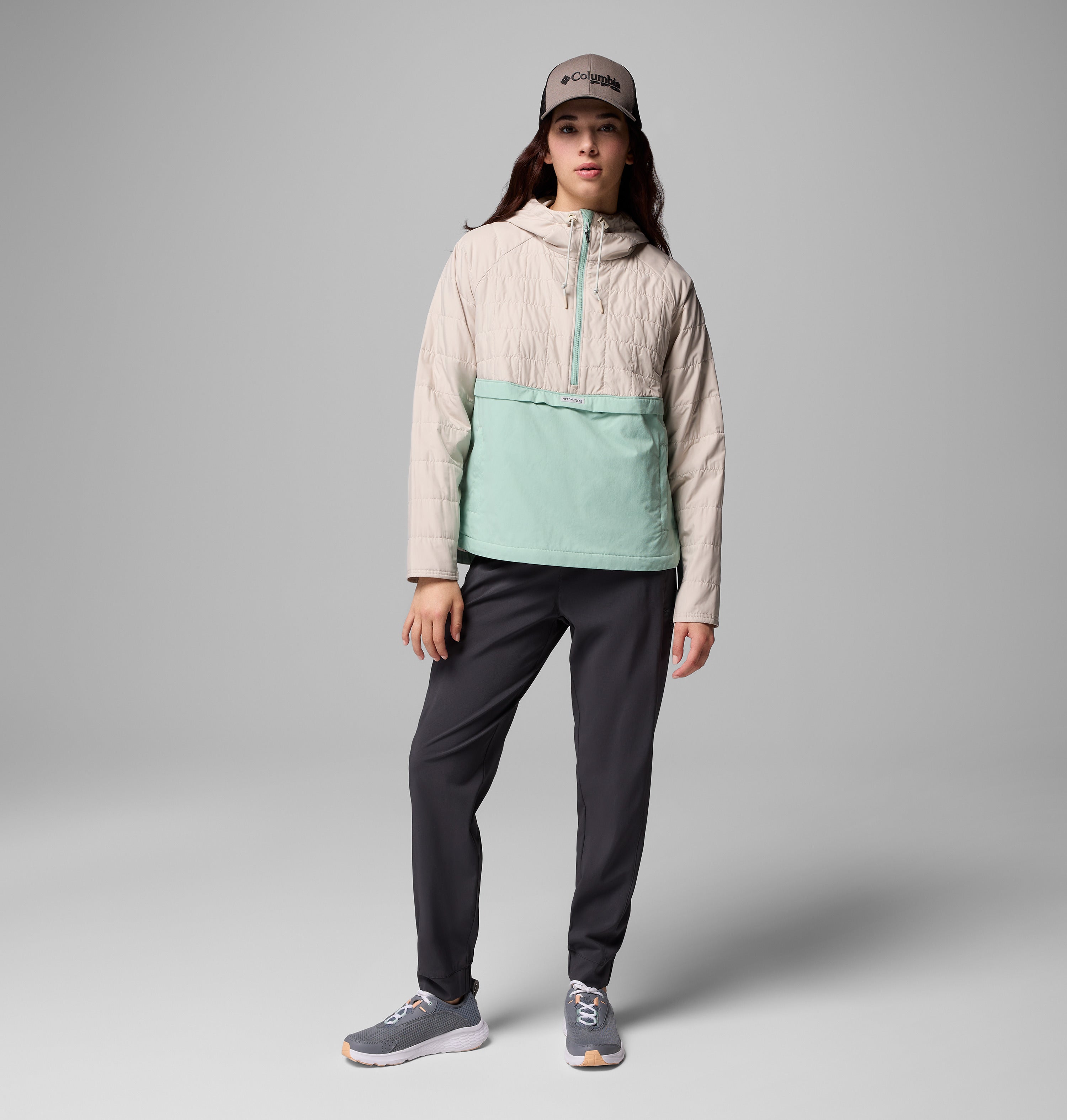 אדם לובש מעיל קולומביה ישראל PFG Uncharted II Insulated Anorak בצבעי בז' וירוק מנטה, מכנסיים כהים, נעלי ספורט אפורות וכובע אפור, בעודו עומד על רקע אפור אחיד.