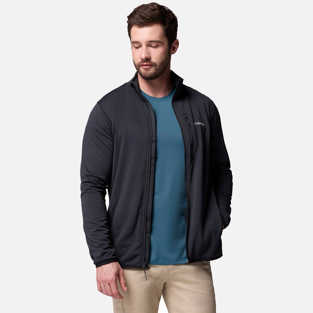 גבר עם שיער חום קצר וזקן לובש את מעיל ה-Essential Hike Grid Full Zip M של קולומביה ישראל מעל חולצה בצבע טורקיז ומכנסיים בצבע בז', עומד עם יד אחת בכיס על רקע לבן חלק.