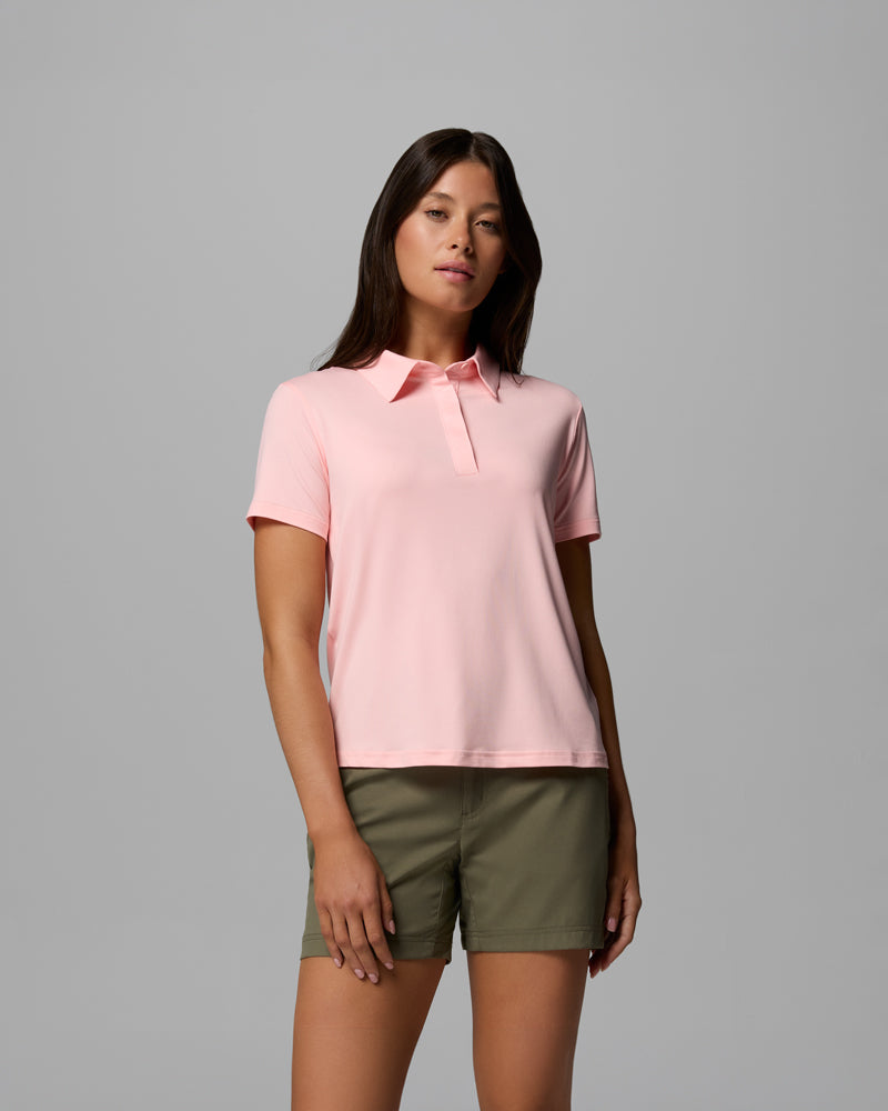 A woman with long brown hair and a neutral expression stands against a plain gray background, wearing the Parsons Point Polo W in light pink and olive green shorts, with her arms relaxed by her sides.