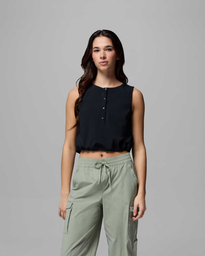 אישה עם שיער חום ארוך לובשת את גופיית ה-Chill Creek Woven Tank של קולומביה ישראל ומכנסי קרגו בצבע ירוק בהיר; היא עומדת על רקע אפור אחיד, פונה קדימה עם הבעה ניטרלית, בסגנון הקלאסי של Chill Creek.