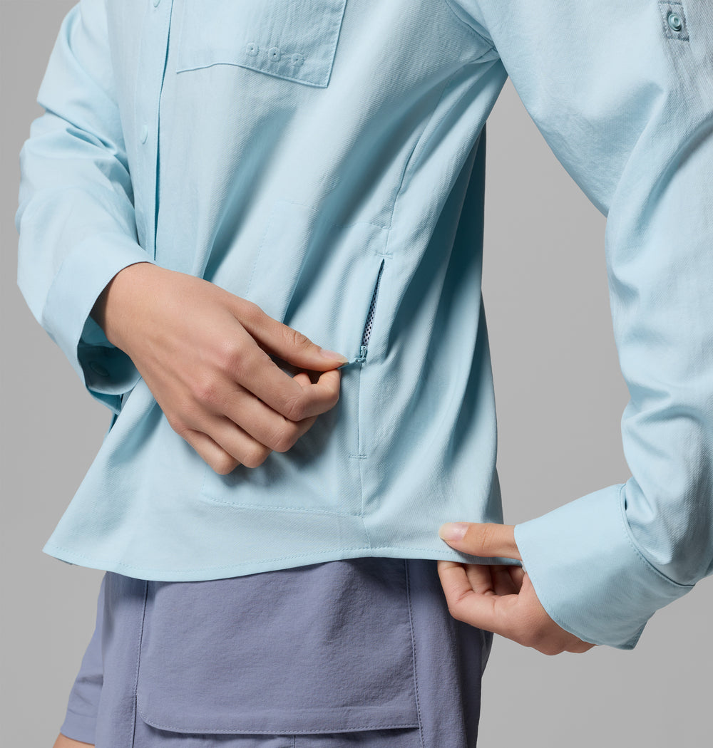 אדם לבוש בחולצת קולומביה ישראל PFG Castback Woven L/S בצבע תכלת ובמכנסיים קצרים בצבע לבנדר פותח את רוכסן הכיס הצדדי בחולצה, על רקע אפור אחיד.