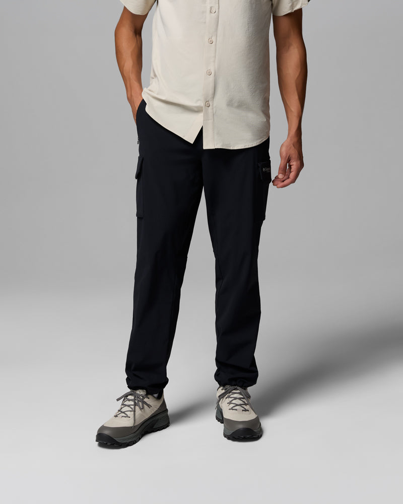 אדם עומד על רקע אפור אחיד, לבוש בחולצה בצבע בז' עם שרוולים קצרים וכפתורים, מכנסי קולומביה ישראל Silver Ridge Elite בצבע שחור, ונעלי טיולים בצבע אפור בהיר. ראשו אינו נראה בתמונה.