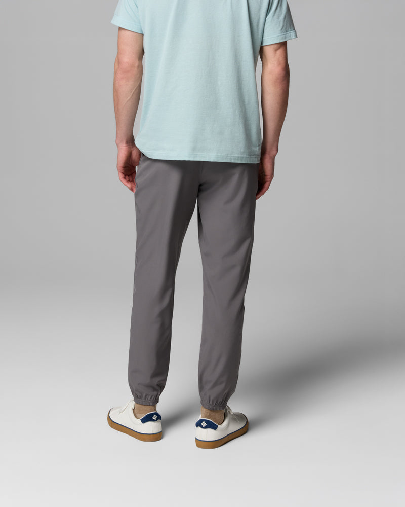 אדם עומד עם גבו למצלמה, לבוש במכנסי ה-Vital Valley Jogger של קולומביה ישראל בצבע אפור, חולצת טי-שירט בצבע תכלת ונעלי ספורט לבנות עם פרטים בכחול, על רקע אפור אחיד.
