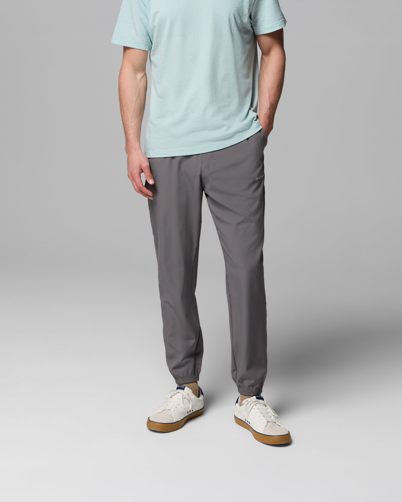 אדם הלובש מכנסי ריצה מדגם Vital Valley Jogger של קולומביה ישראל בצבע אפור, חולצת טי עם שרוולים קצרים בצבע תכלת ונעלי ספורט לבנות, עומד כש יד אחת בכיס על רקע אפור אחיד.