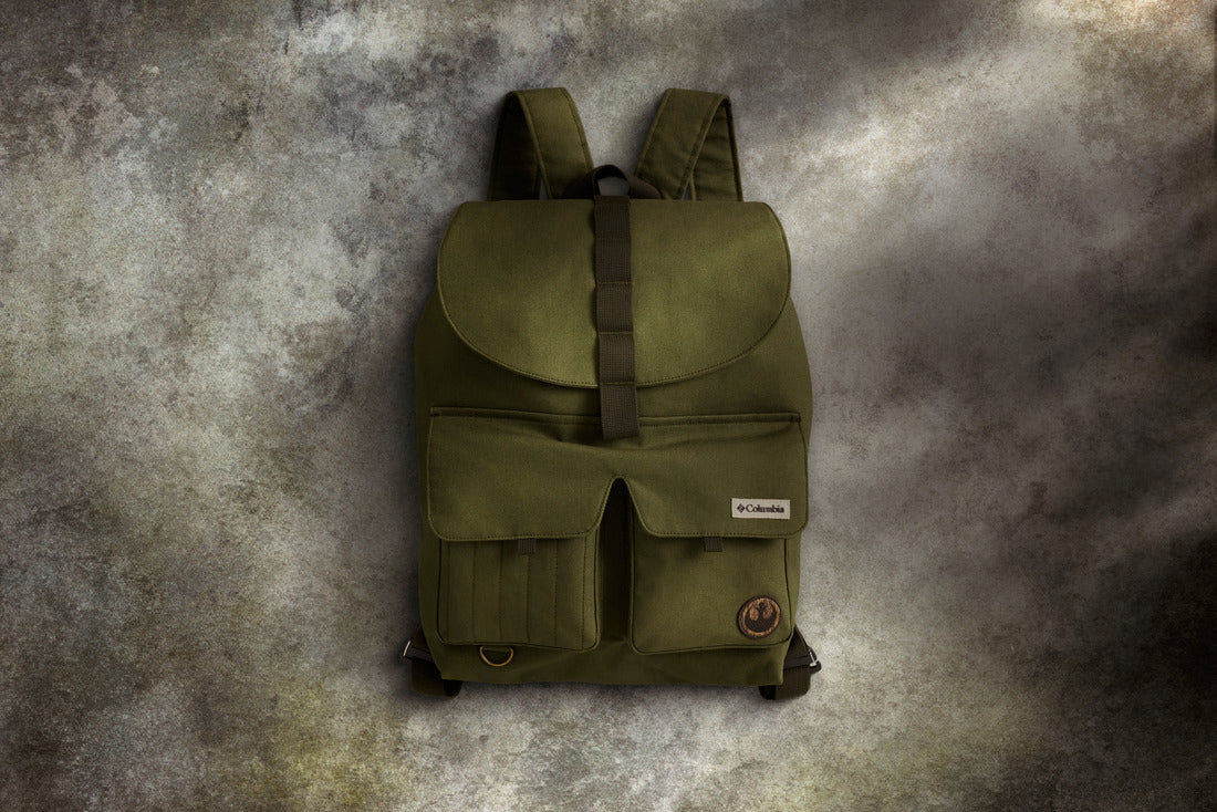 תיק הגב "Endor Issue Cargo" של קולומביה ישראל בצבע ירוק זית כולל שני כיסים קדמיים, רצועה חומה ותווית עגולה על הכיס הימני התחתון, המוצגת על רקע אפור מרקם.